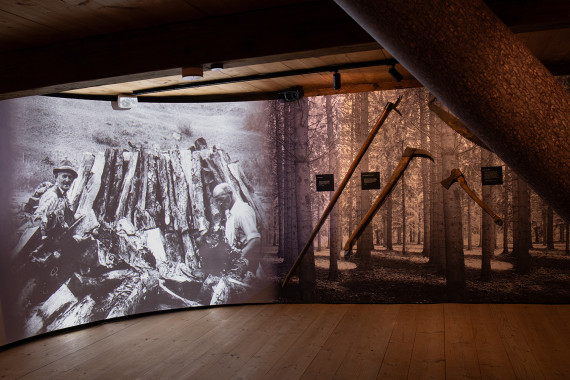 Holzraum der Ausstellung im Blaahaus mit gebogener Videoleinwand Werkzeugen, ein fallender Baumstamm schneidet das Bild an.