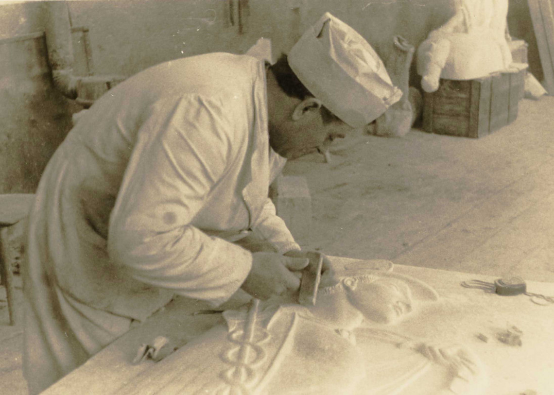 Ein Marmorsteinmetz mit Papierkappe ist über eine Marmorplatte gebeugt und meiselt an einem Relief