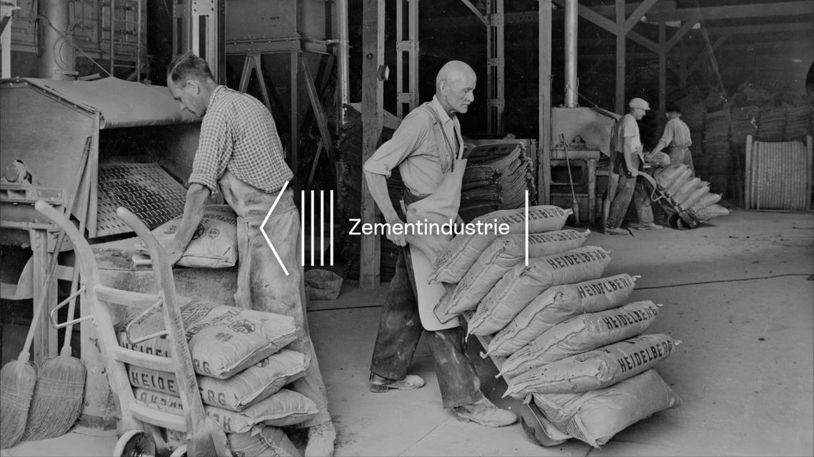 This black-and-white photograph shows emaciated forced labourers stacking sacks of cement onto sack trucks and removing them from conveyor belts in a production hall. Each sack weighs 50 kg.