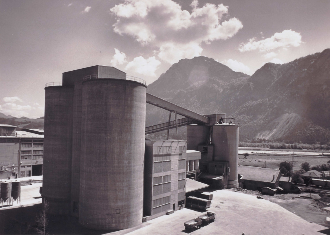 Zwei hohe Betonsilos und verschiedene andere Bauwerke der Zementproduktionsstätte in Kiefersfelden, dargestellt in Schwarz-Weiß.