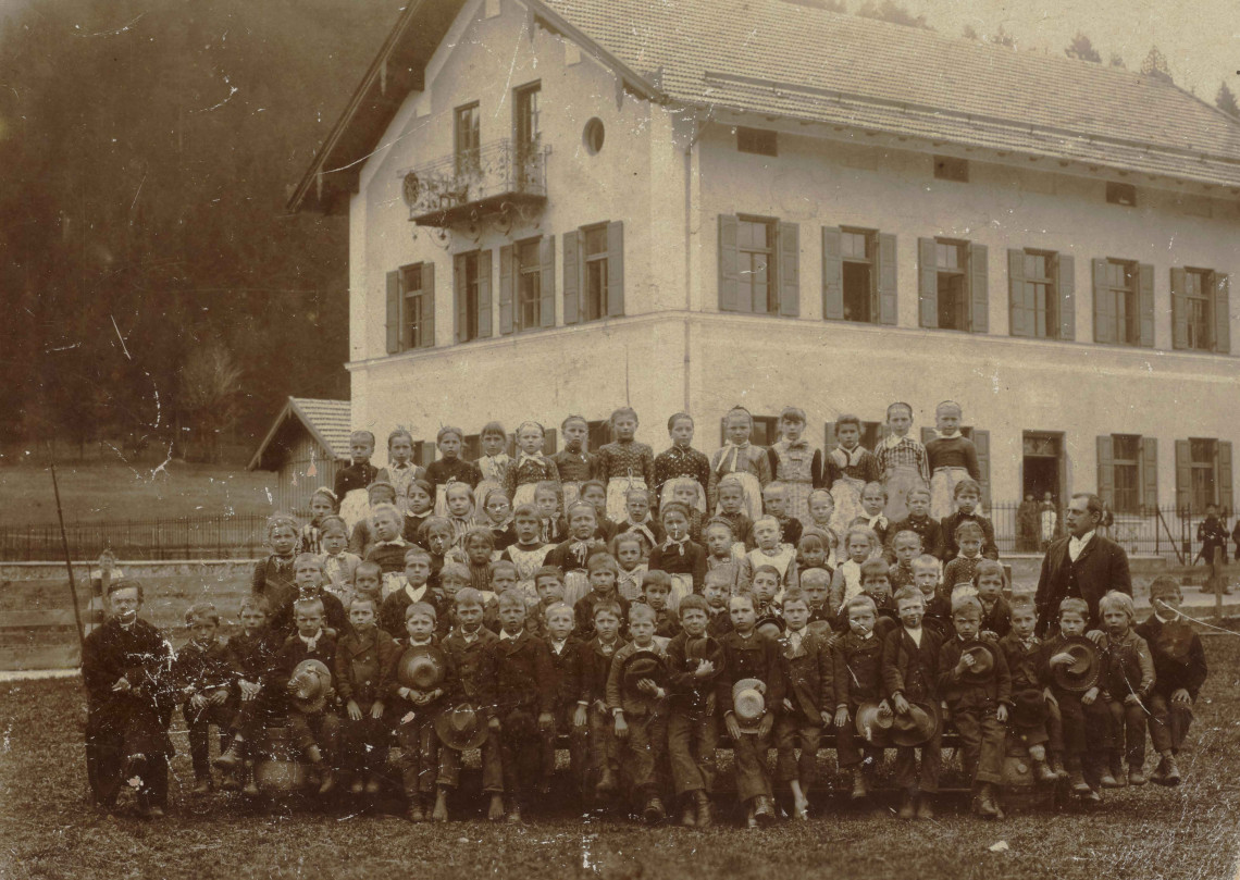 Ein sepiafarbenes Gruppenfoto, das mindestens 78 Kinder und zwei männliche Lehrer zeigt, die vor einem neu-errichteten ländlichen, dreistöckigen Schulgebäude stehen.