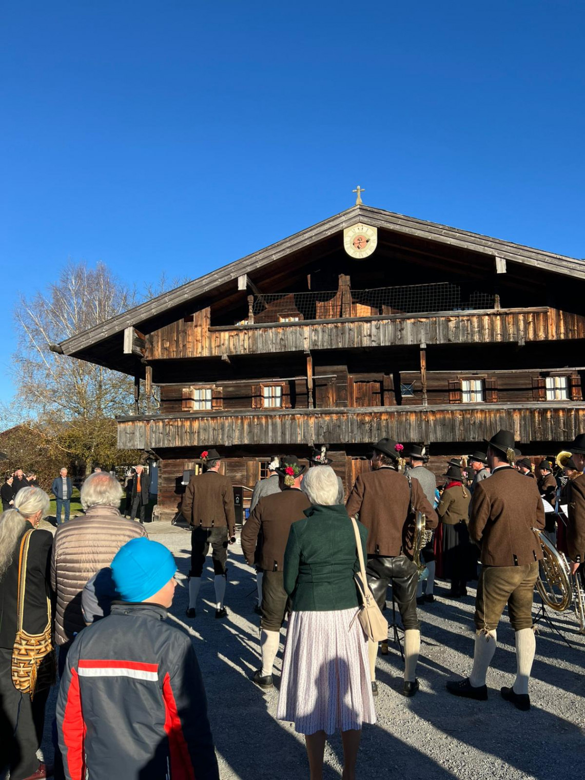 Auf dem Bild steht das neueröffnete Industriemuseum Blaahaus Kiefersfelden vor strahlend blauem Himmel. Davor sieht man auf dem Kiesplatz von hinten die große Gruppe an Gäste und Musikanten in ortstypischer Tracht.
